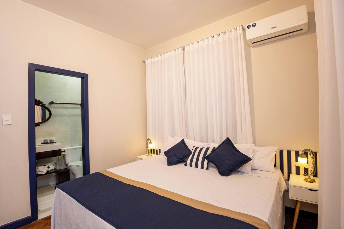 A hotel room with a bed, bedside table, nightstand, and an air conditioning unit.