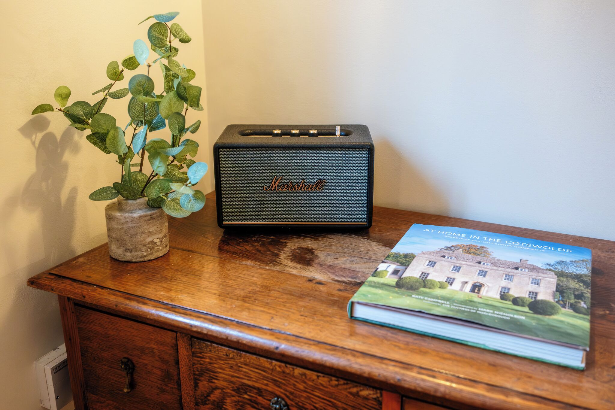 Un escritorio de madera con un altavoz Marshall, una planta en maceta y un libro titulado 'At Home in the Cotswolds'.