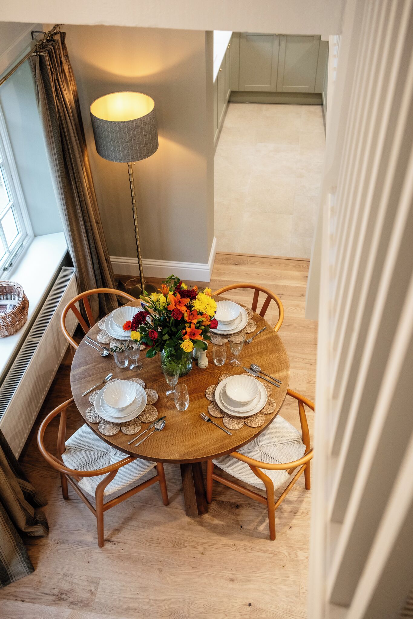 A dining area with a round table set for four, surrounded by wooden chairs. A lamp is positioned to the left of the table, and a window with curtains is visible in the background.