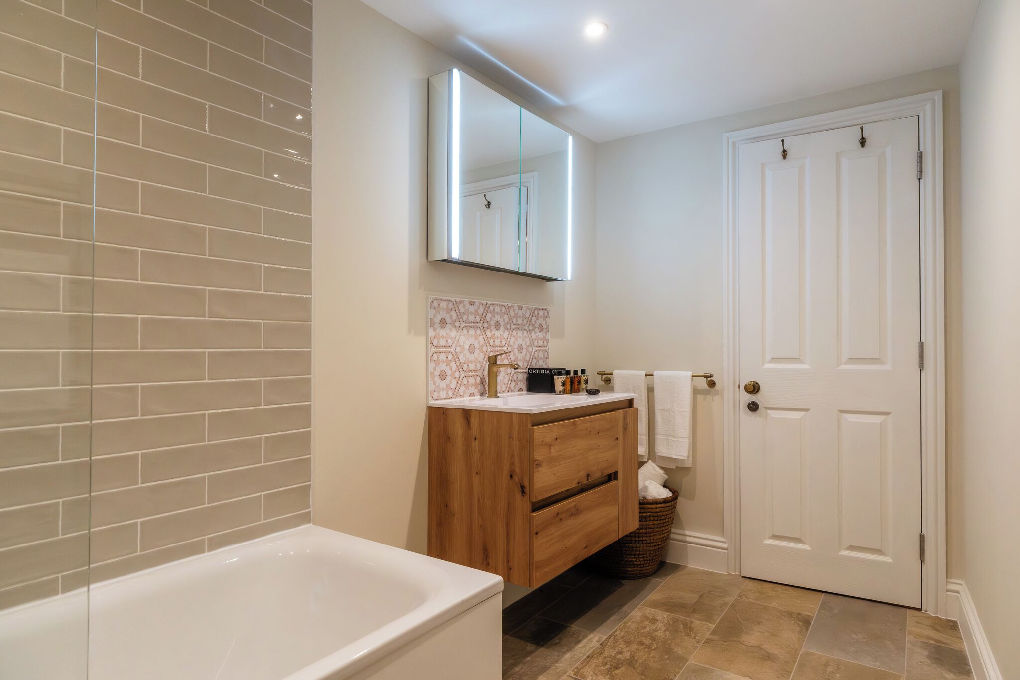 A bathroom with a white bathtub, a wooden vanity with a mirror, and a white door.