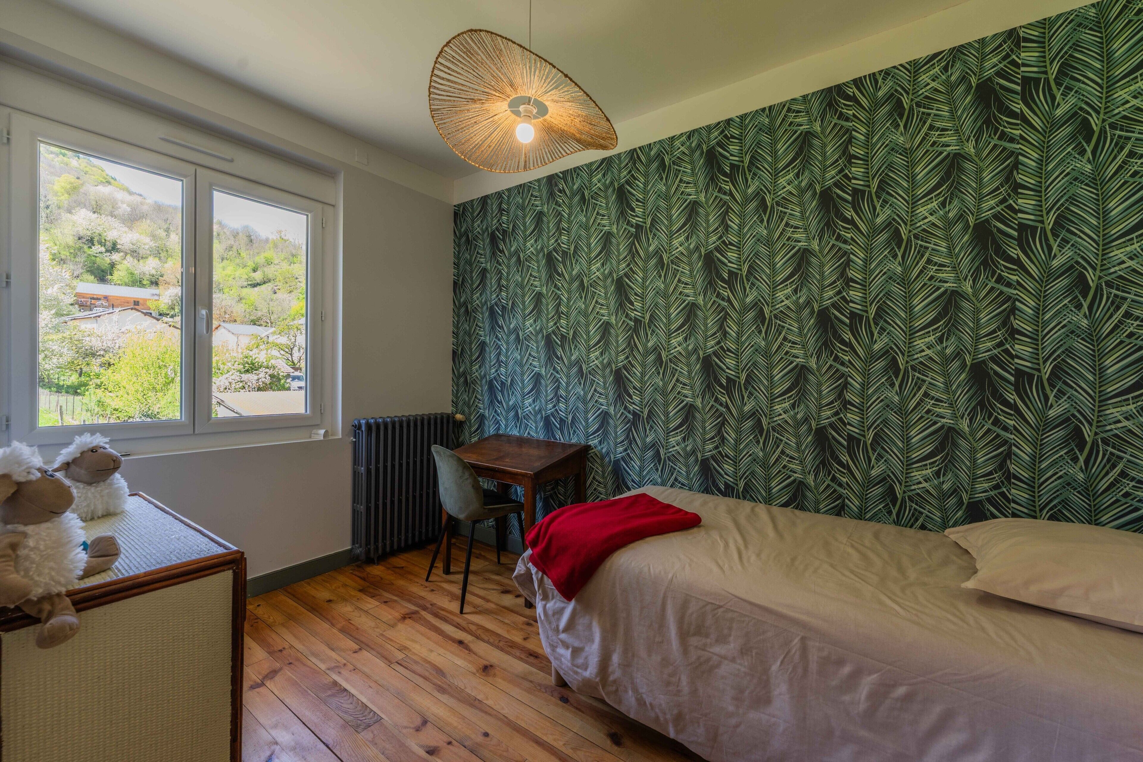 Een slaapkamer met een bed, een bureau, een stoel, een radiator en een raam met uitzicht op groen.
