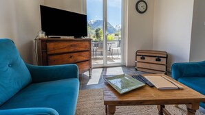 A living room with a TV, two blue sofas, a wooden table with a laptop and a notebook, a wooden chest, and a sliding glass door leading to a balcony with a mountain view.