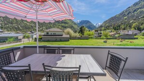 A patio with a table and chairs, a striped umbrella, and a view of a green landscape with houses and mountains.