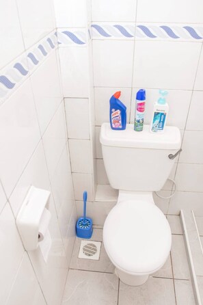 A bathroom with a toilet, a blue toilet brush, a paper towel dispenser, and three toiletry bottles on the tank. - Mora studio in Nyali Oshwal Academy 4B (Mombasa)