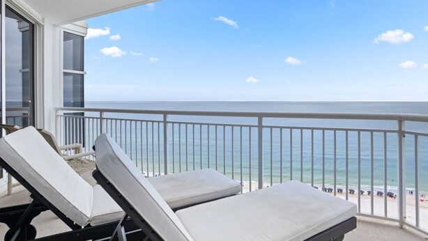 Ein Balkon mit zwei Liegestühlen, die zum Meer hinweisen, einem weißen Geländer und freiem Blick auf den Strand.