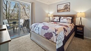 A bedroom with a bed, two bedside lamps, a ceiling fan, a window with a view of trees, and a small table with chairs. - NEWLY RENOVATED COZY 2BR 1BA COTTAGE -NEW HOT TUB (Sautee Nacoochee)