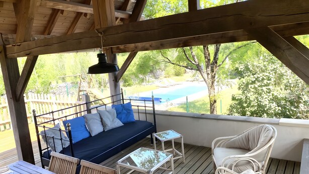 Een overdekt terras met een bank, twee tafels en stoelen, met uitzicht op een zwembad en groen.