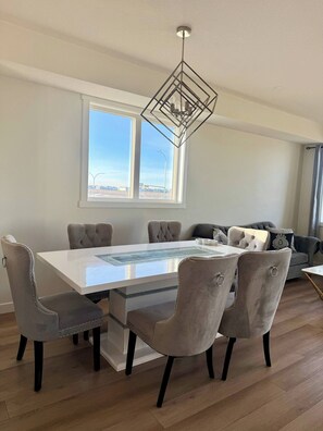 A dining area with a white table, grey upholstered chairs, a geometric ceiling light, and a view of the outdoors through large windows. - Modern Living - Corner Unit Townhome 3 Bedrooms (Calgary)