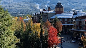 Un paese di montagna con un edificio importante caratterizzato da un campanile, circondato da alberi dai colori autunnali e uno sfondo di montagne innevate.