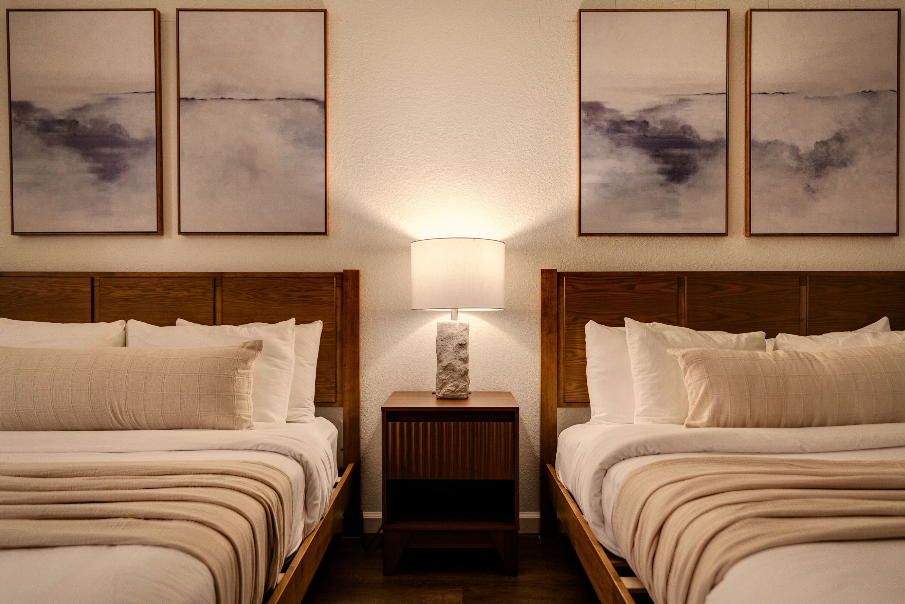 Une chambre d’hôtel avec deux lits, une table de chevet avec une lampe et trois tableaux abstraits accrochés au mur.