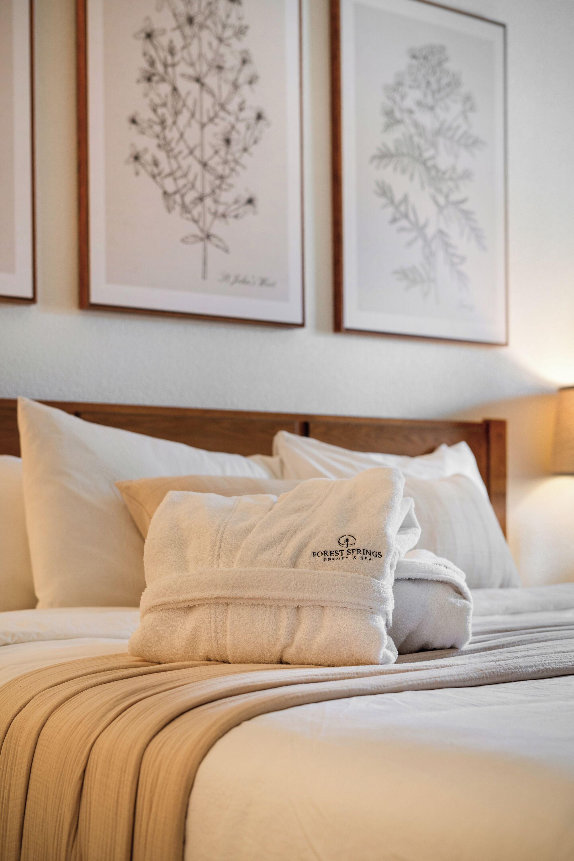 A neatly made bed with white linens, a beige throw blanket, and a Forest Springs branded bathrobe.