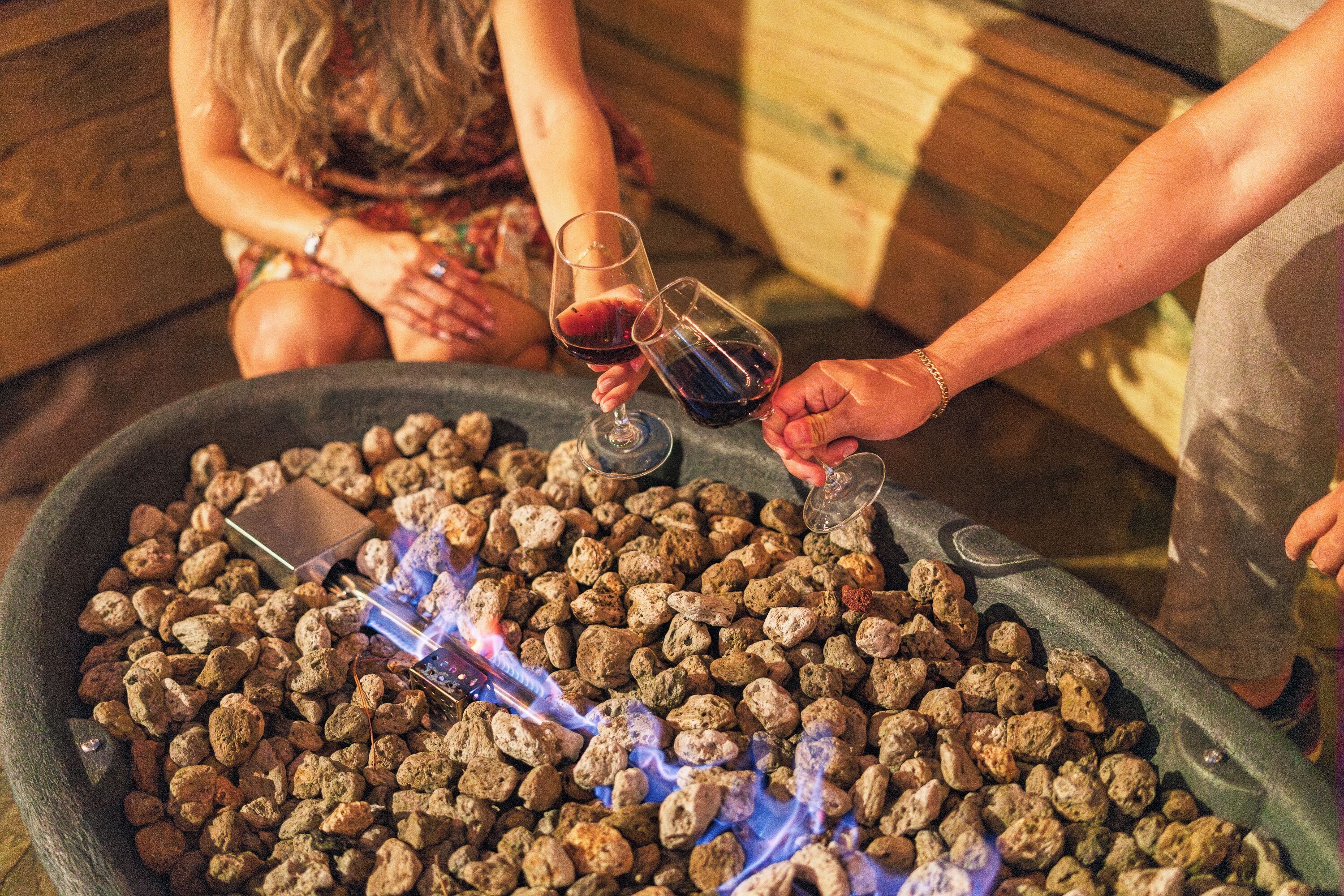 A large stone fire pit with rocks and a visible flame, surrounded by people.
