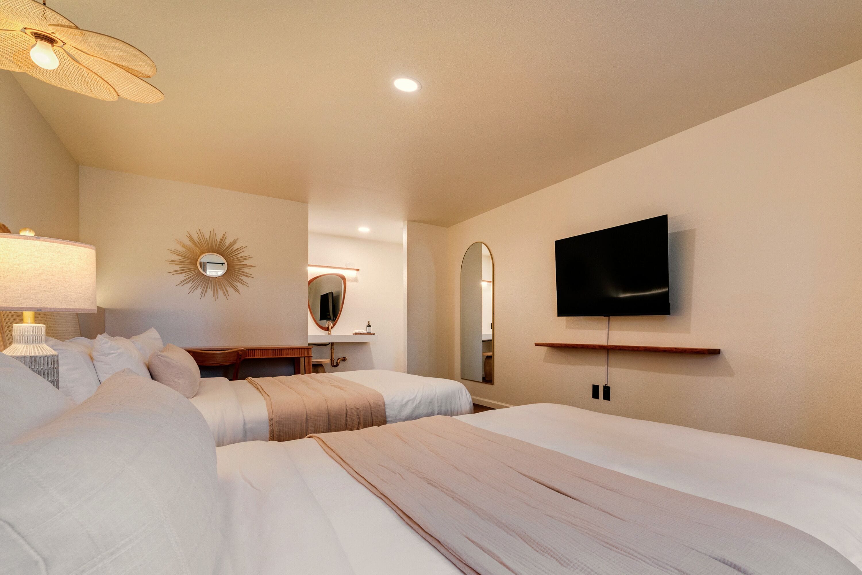 A hotel room with two beds, a television mounted on the wall, a ceiling fan, and a mirror.
