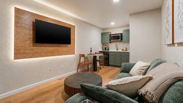 Una sala de estar moderna con un televisor de pantalla plana montado en un panel de madera, un sofá verde con una bandeja de metal y una zona de comedor con una mesa y sillas.