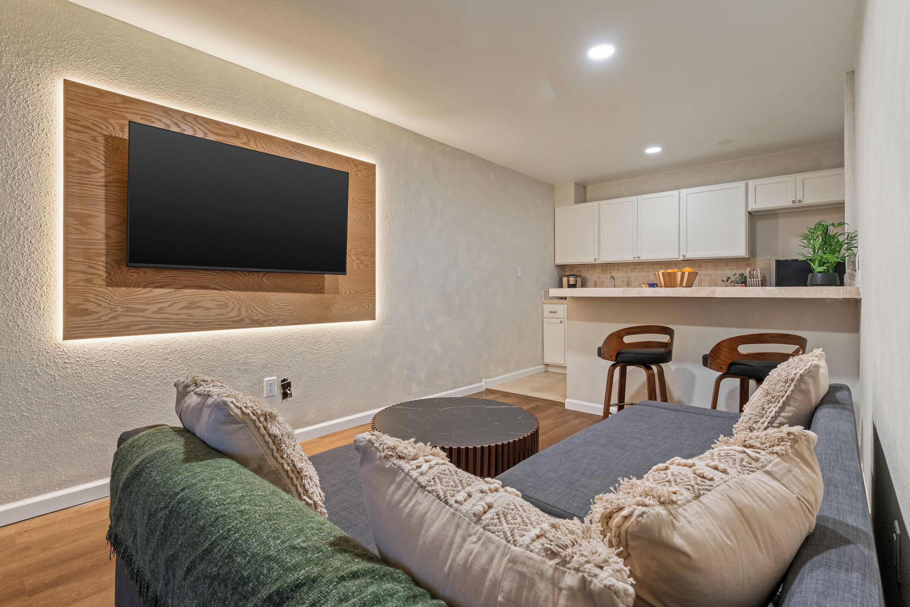 Una sala de estar moderna con un televisor de pantalla plana montado en una pared texturizada, una mesa de centro redonda y un sofá con almohadones decorativos.