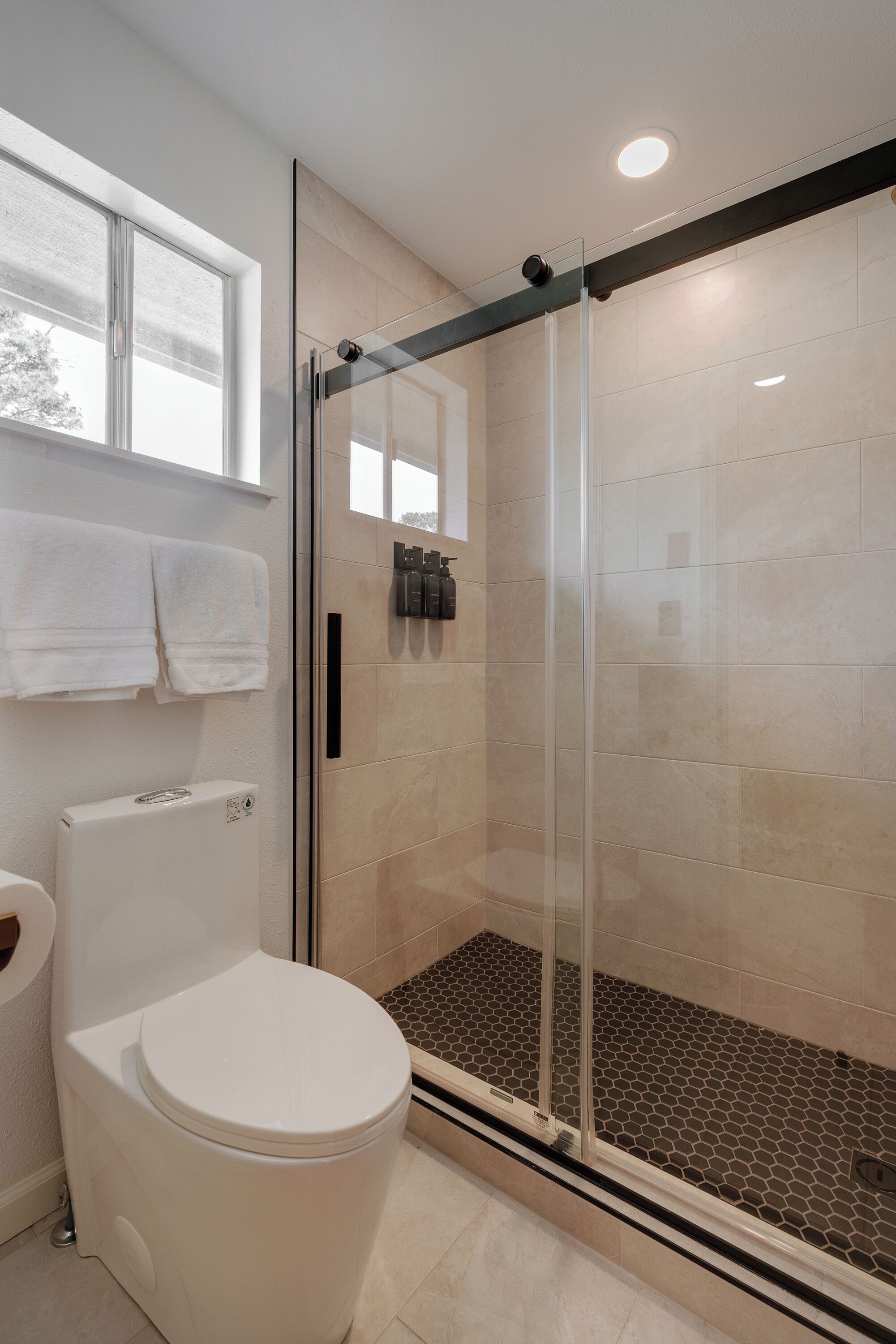 A modern bathroom with a glass-enclosed shower, a toilet, and a window above the sink.