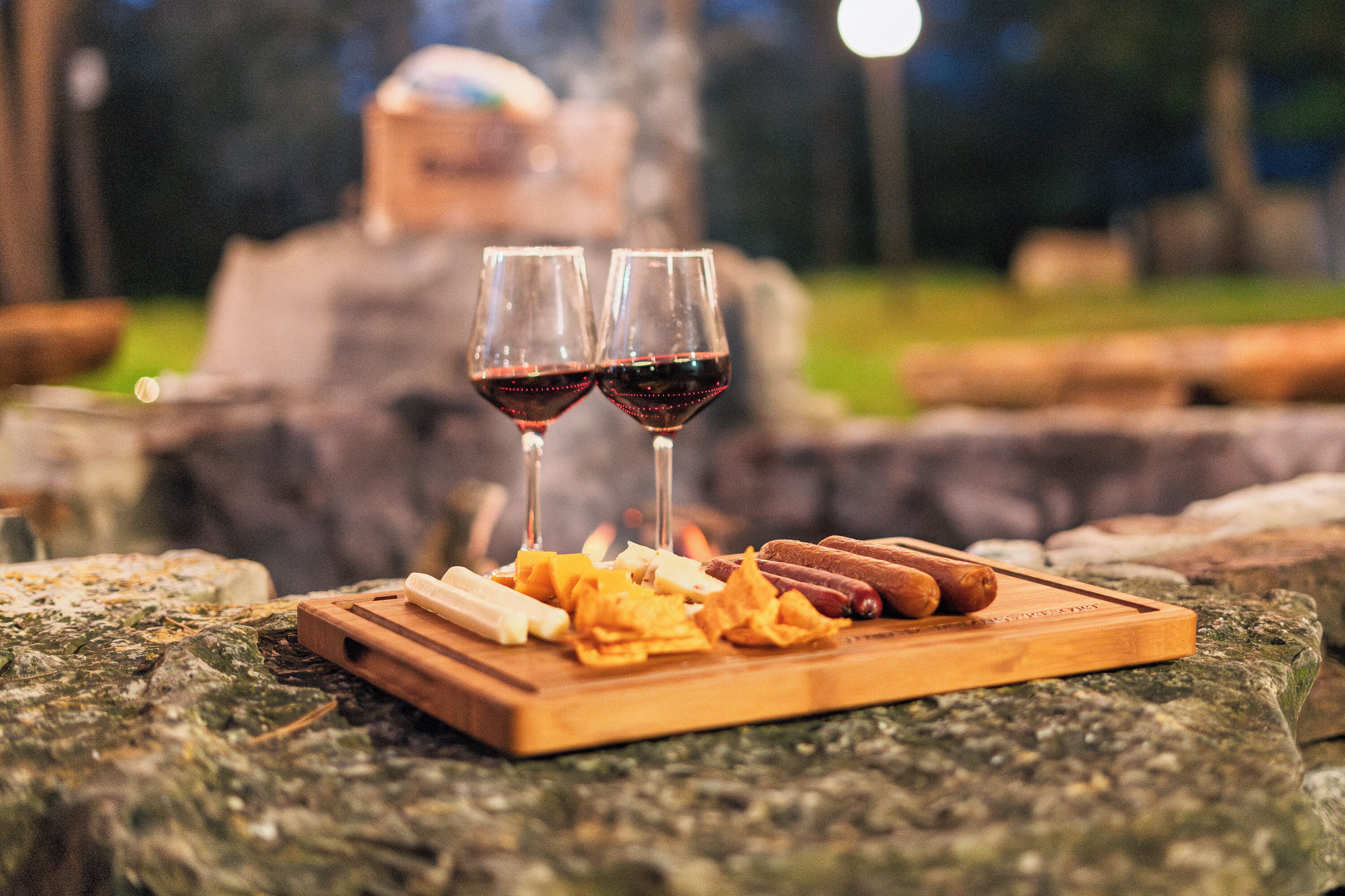 A wooden cutting board with snacks and two glasses of red wine on a stone surface.