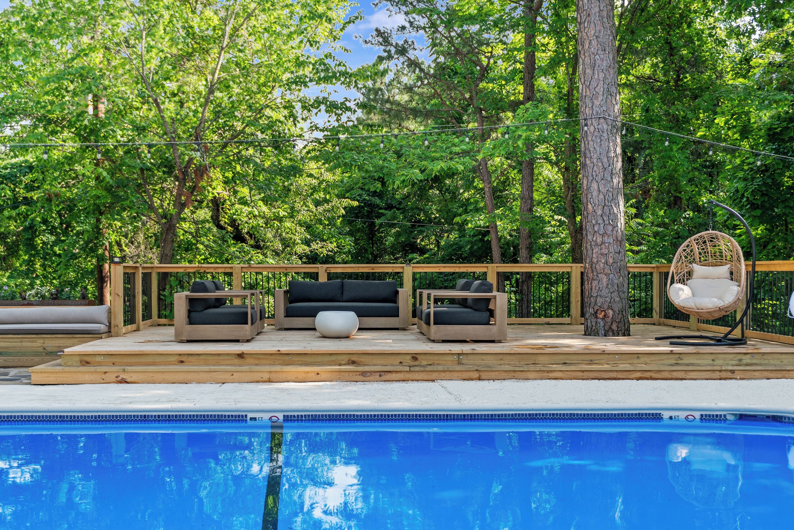 A poolside area with outdoor furniture, a wooden deck, and a view of trees.