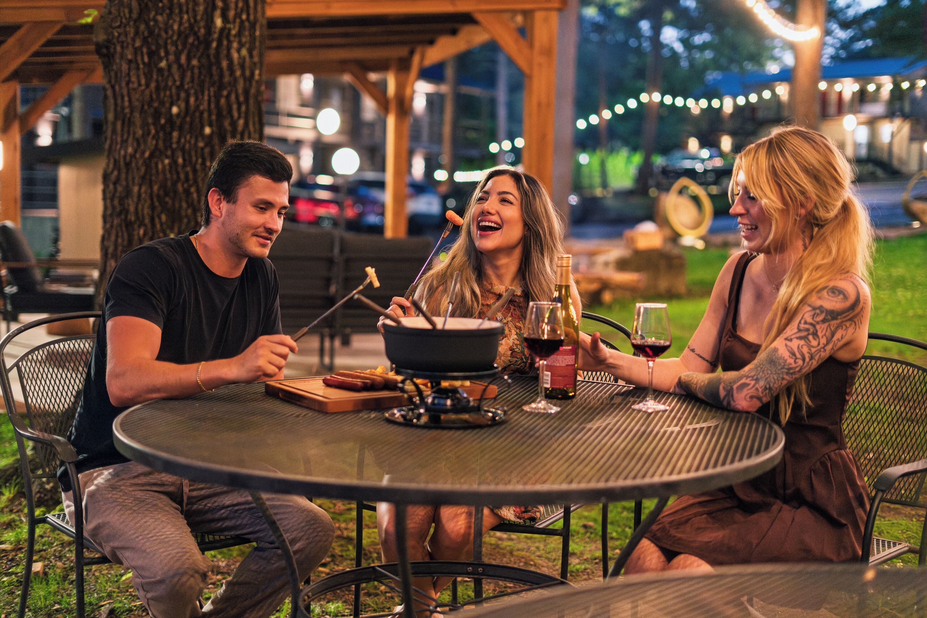 Three people dining outdoors at a round metal table with a pot and wine glasses.