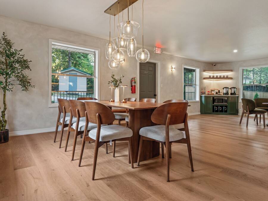 Un comedor amplio con una mesa y sillas de madera, una planta grande en maceta y vista hacia una casa a través del ventanal.