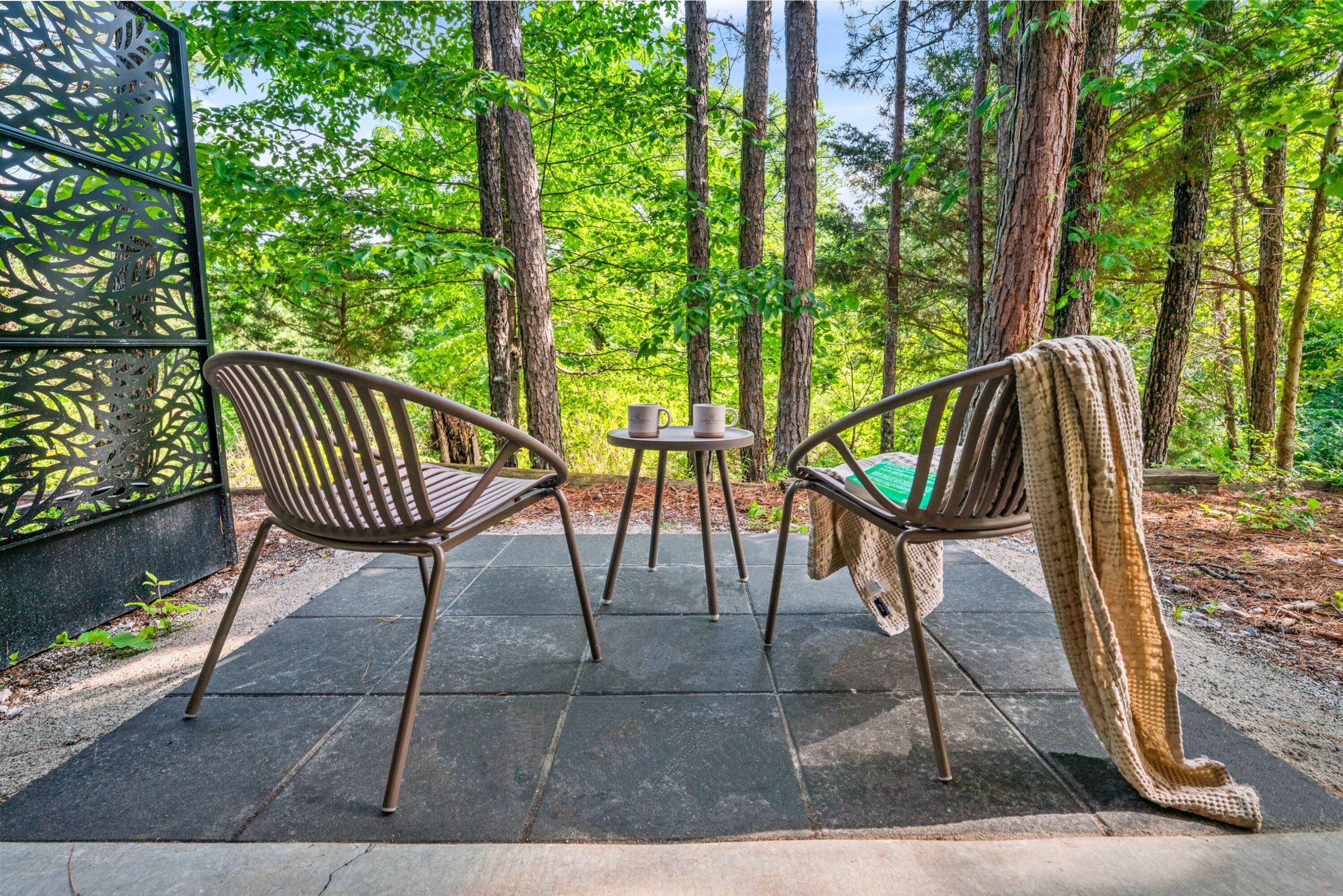 A patio with two chairs and a table, surrounded by a forest.