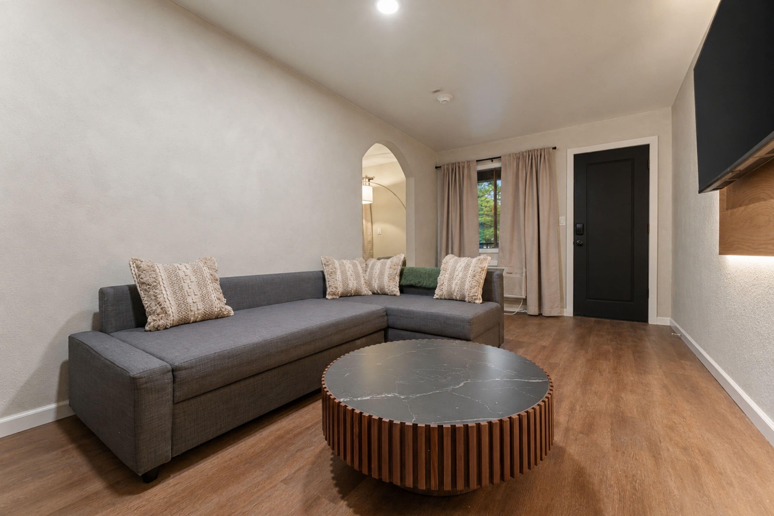 Un salon moderne avec un canapé d’angle, une table basse ronde, un parquet en bois et une porte noire.
