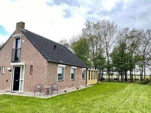 A brick house with a sloped roof, a door, and windows. There is a lawn with a few chairs and a table. - Beautiful Cottage for 6 in Ewijk Park (Ewijk)