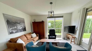 A living room with a sofa, two armchairs, a coffee table, a TV, and a large window with a view of greenery. - Beautiful Cottage for 6 in Ewijk Park (Ewijk)