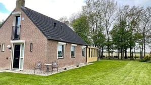 A brick house with a sloped roof, a door, and a small patio with two chairs. - Beautiful Cottage for 6 in Ewijk Park (Ewijk)