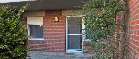 Un edificio residencial con fachada de ladrillo, un pequeño jardín delantero con césped, una rejilla metálica y una ventana con persiana blanca.