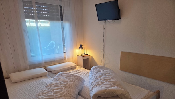 Ein Hotelzimmer mit Bett, Kissen, Nachttischlampe, Fernseher und einem Fenster mit Jalousien.