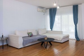 Un salon comprenant un canapé, une table basse, une table d’appoint et une fenêtre avec des rideaux.