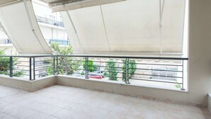 A balcony with a retractable awning, tiled floor, and a view of a red car and greenery. - Bright Minimal 2 Bd Apt in Marousi (Marousi)
