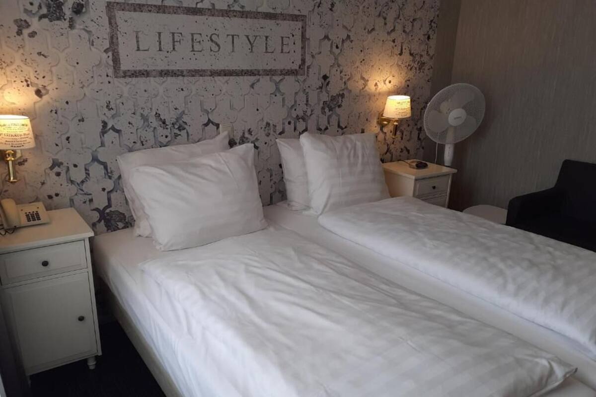 Una cama doble bien hecha con ropa de cama blanca, dos mesitas de noche con lámparas, un reloj de pared y un cartel enmarcado que dice 'LIFESTYLE'.