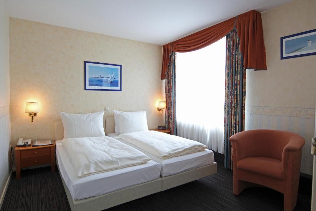 Een hotelkamer met een tweepersoonsbed, twee nachtkastjes met lampen, een stoel, een raam met gordijnen en een ingelijst schilderij aan de muur.