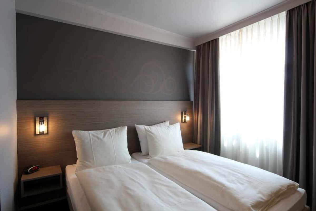 Una habitación de hotel con una cama grande, dos mesitas de noche con lámparas, un cabecero con luz y una ventana con cortinas.
