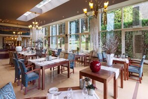 A spacious dining area with multiple tables and chairs, large windows offering a view of greenery, and elegant chandeliers. - Grand Hotel Monopole (Valkenburg aan de Geul)