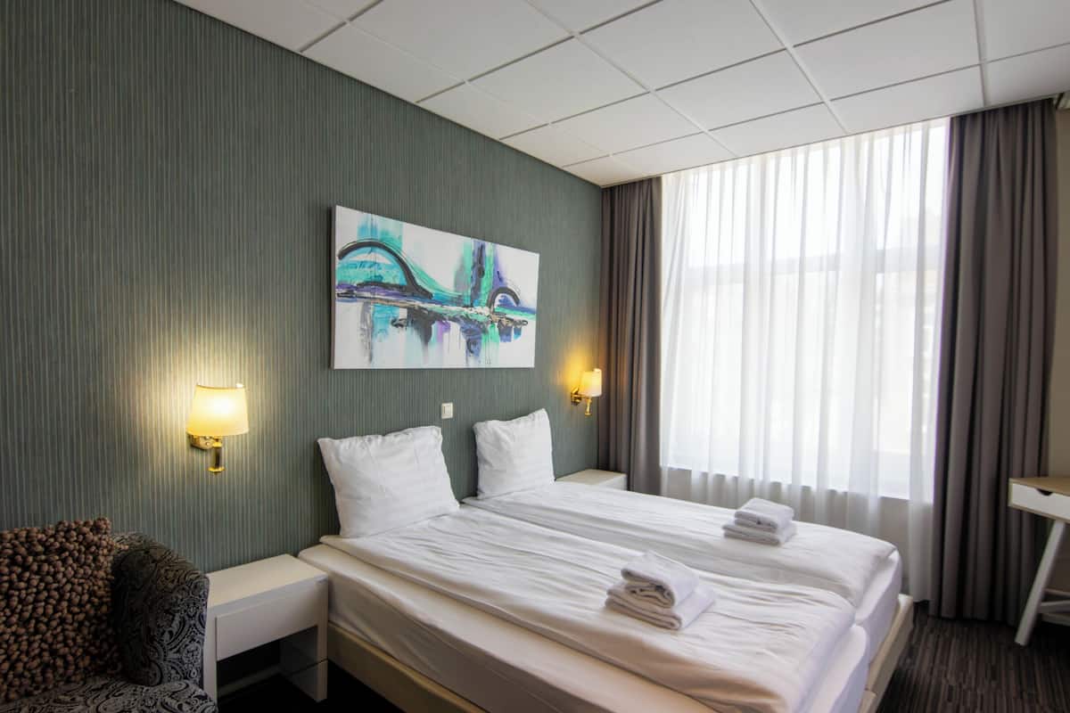 Habitación de hotel con una cama, dos mesitas de noche, un cuadro colgado en la pared y una ventana con cortinas.