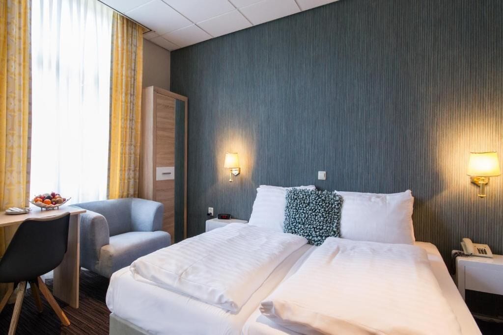 A hotel room with a bed, a desk, a chair, a bedside table with a lamp, and a telephone.