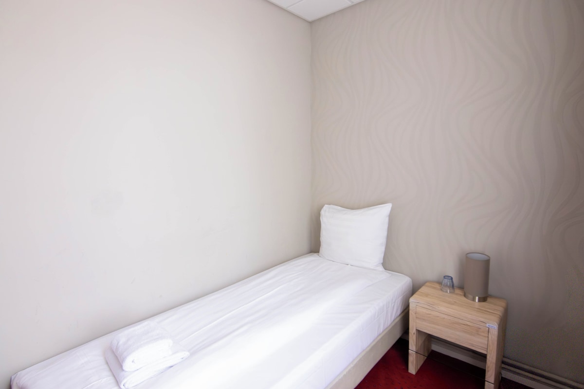 Una cama individual con colchón y almohada blancos, una mesita de noche de madera con lámpara y una pared con textura.