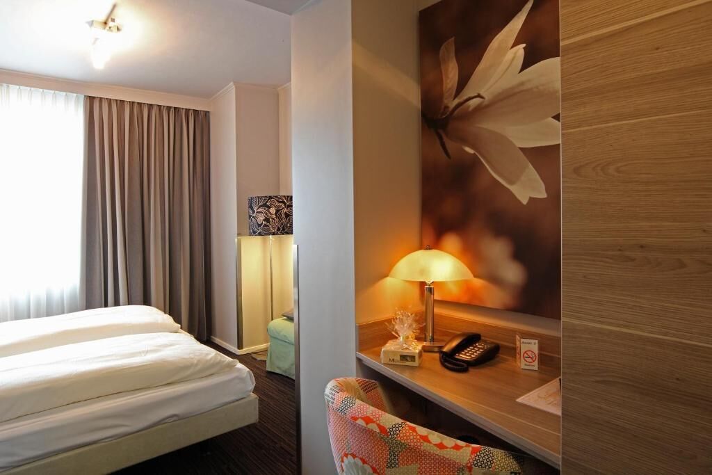 Habitación de hotel con cama, escritorio con lámpara, teléfono y un cuadro floral en la pared.