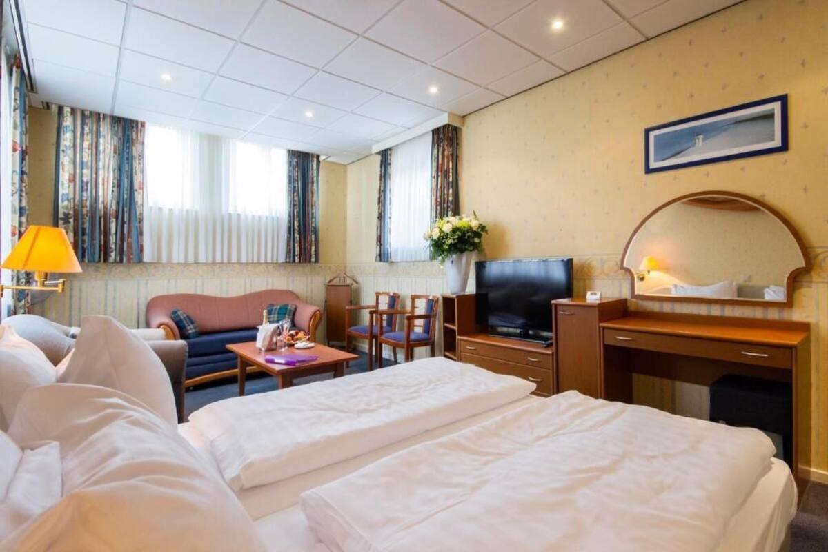 Habitación de hotel con dos camas, televisor, escritorio, silla, lámpara y ventana con cortinas.