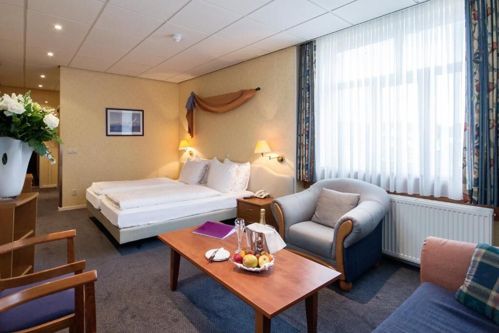 A hotel room with a bed, two bedside tables, a chair, a sofa, a coffee table with fruit, and a window with curtains.