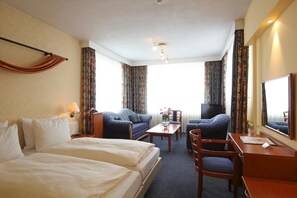 A hotel room with two beds, a desk, a chair, a TV, and curtains. - Grand Hotel Monopole (Valkenburg aan de Geul)
