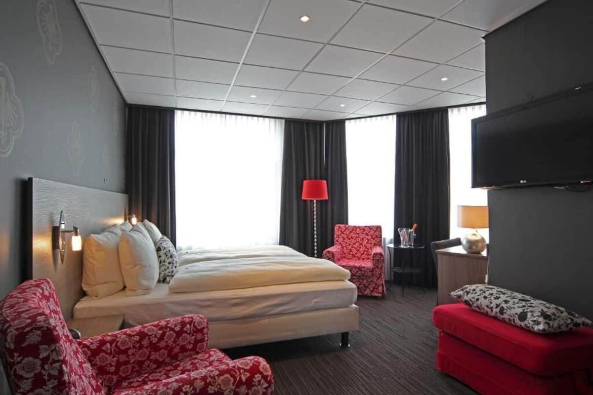 Una habitación de hotel con una cama, un sillón rojo, un sofá rojo, un escritorio con lámpara y un televisor.