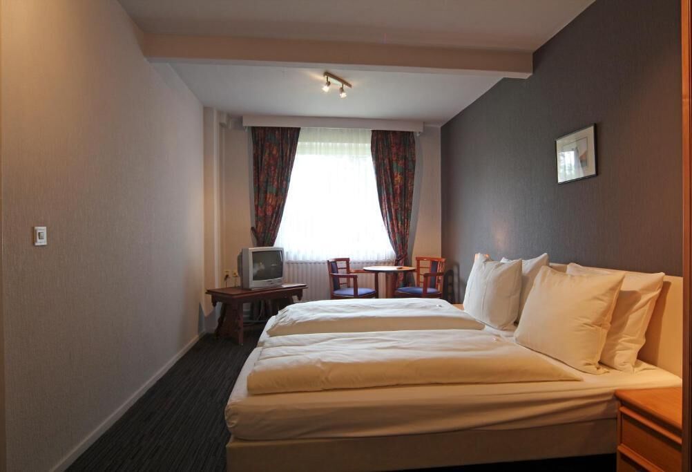 A hotel room with a double bed, a television, a small table, two chairs, and a window with curtains.
