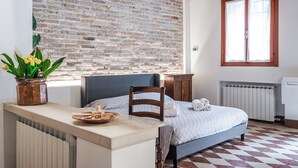 Ein Zimmer mit Backsteinwand, einem Bett mit blauer Kopfteil, einem Holzsessel, einem kleinen Tisch mit Tablett und einem Fenster mit Holzrahmen.