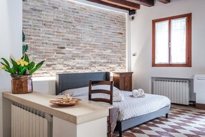 A room with a brick wall, a bed with a blue headboard, a wooden chair, a small table with a tray, and a window with a wooden frame. - GDL HOUSE VENICE (Venezia)