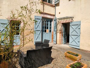 Een binnentuin met rieten meubilair, een houten plantenbak met bloemen en blauwe luiken aan het gebouw.