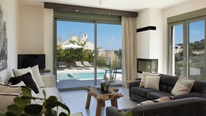 Ein modernes Wohnzimmer mit einer Couch, einem Couchtisch und einem Fernseher. Ein großes Fenster bietet Blick auf einen Pool und viel Grün außerhalb.